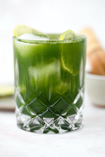 A frosty green matcha cocktail in a clear, patterned glass garnished with lime slices. The background features a blurred whisk and small bowl. Refreshing and inviting.