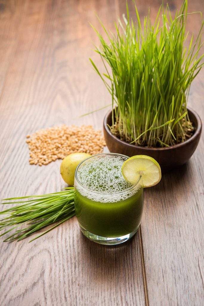 A refreshing green juice garnished with a slice of lime, served alongside wheatgrass and grains on a wooden surface.